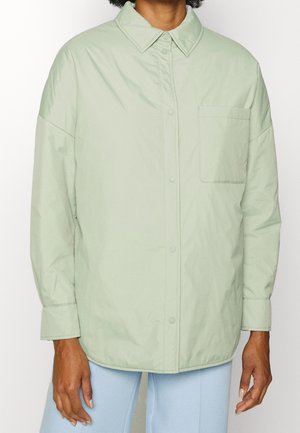 Person wearing a light green button-up shirt with front pocket and light blue pants, standing against a plain background.