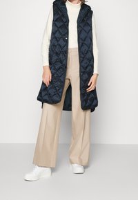 Chaleco acolchado navy con capucha, que presenta patrones de diamante y cierres a presión, combinado con pantalones de pierna ancha beige y zapatillas blancas.