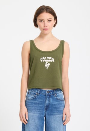 Jeune femme portant un débardeur court vert olive avec le texte "Volcom" et un motif floral, associé à un jean taille haute bleu.