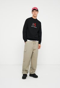 Czarny sweatshirt z kolorową grafiką i napisem, beżowe spodnie cargo z bocznymi kieszeniami oraz różowa czapka. Całość dopełniają czarne obuwie.