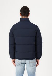 Marineblauwe gewatteerde jas met een hoge kraag, voorzien van horizontale gestikte panelen en elastische manchetten, gecombineerd met lichtblauwe jeans.