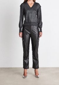 Metallic schwarzer V-Ausschnitt-Pullover mit langen Ärmeln, kombiniert mit schwarzen Fake-Leder-Hosen mit Seitentaschen und Reißverschlussdetails. Silberne Absatzschuhe.