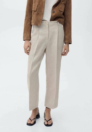 Woman wearing beige high-waist straight trousers, brown cropped jacket, and black open-toe low heel sandals, standing against a plain background.