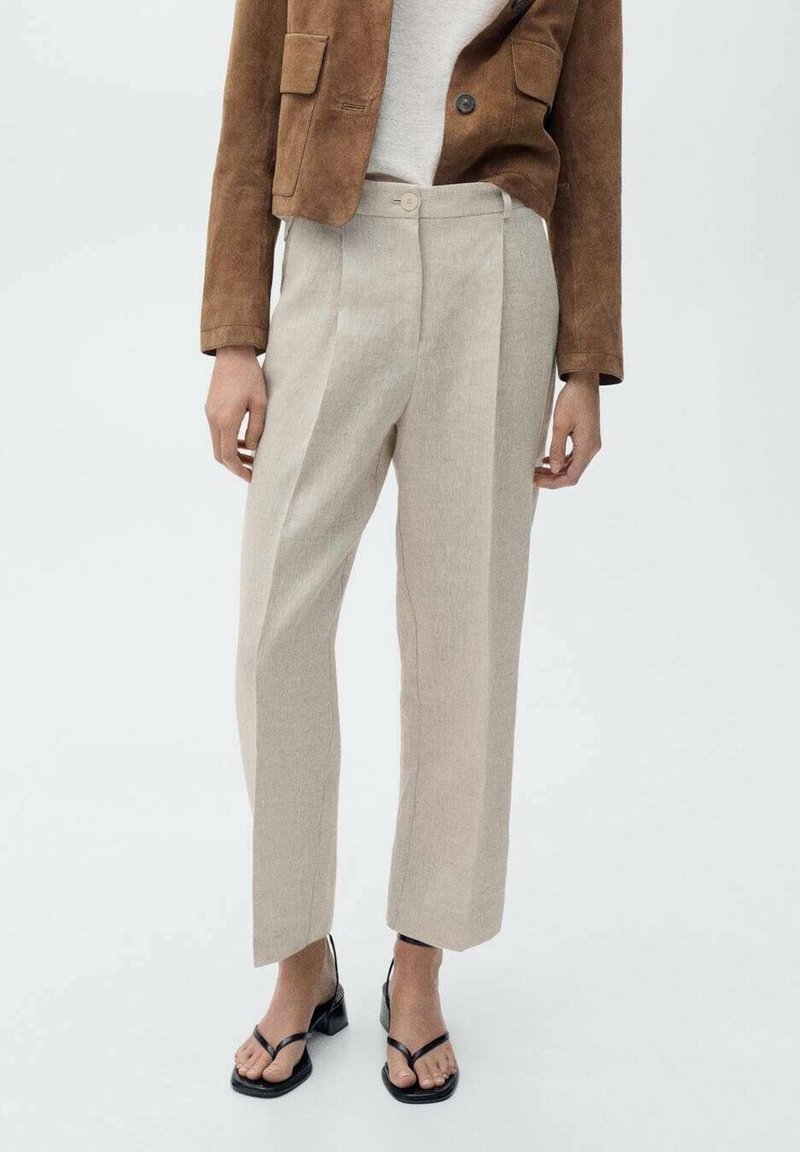 Femme portant un pantalon beige taille haute droit, une veste courte marron et des sandales noires à talon bas et bout ouvert, debout devant un fond uni.