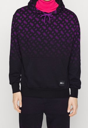 Mann trägt einen schwarzen und lila gemusterten Kapuzenpullover mit einer vorderen Kängurutasche und lilanen Kordeln, darüber einen knallpinkfarbenen Rollkragenpullover.