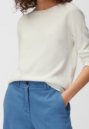 Pull en maille beige clair avec un col rond et des manches trois-quarts, associé à un pantalon bleu avec poches latérales et un bouton.