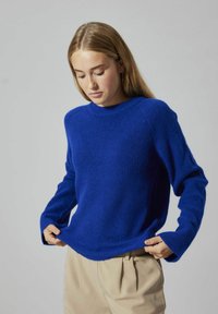 Blauer Strickpullover mit Rundhalsausschnitt und gerippter Textur, mit langen Ärmeln und oversized Passform, kombiniert mit beigen Hosen.