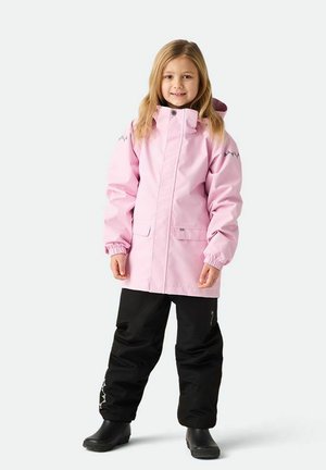 Ung pige smiler, iført en pink vandtæt jakke og sorte skibukser, stående på en hvid baggrund.