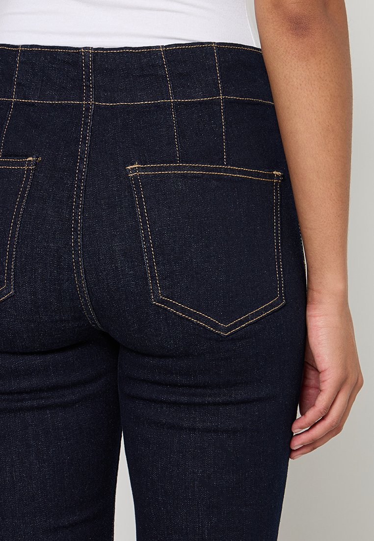 Jeans en denim foncé avec une coupe ajustée, dotés de deux poches arrière, d'une couture jaune contrastée et d'une texture lisse.
