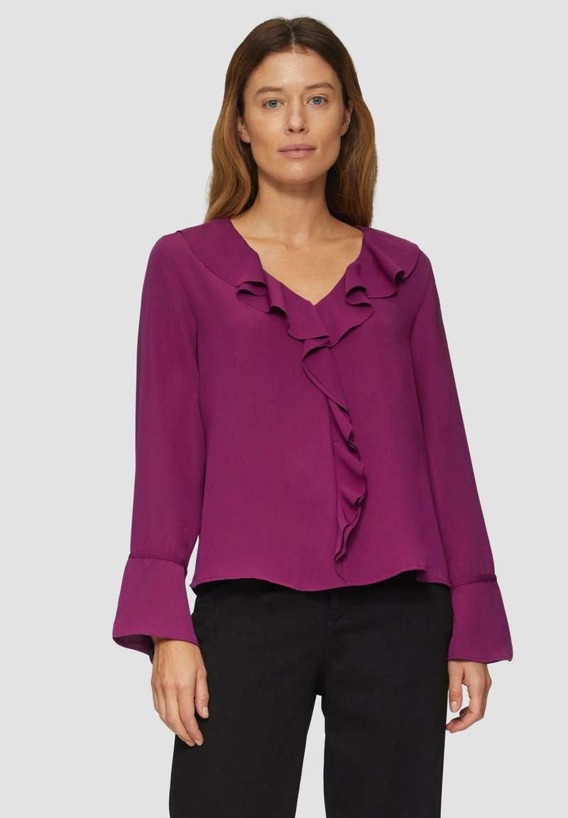Blouse violette en tissu texturé avec un col en V, des volants à l'avant et des manches évasées, associée à un pantalon noir.