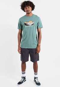 T-shirt en coton turquoise avec imprimé graphique ; associé à un short cargo foncé. Chaussettes blanches avec des rayures noires et baskets montantes noires.