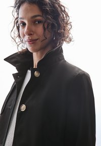 Czarna, dopasowana marynarka z wysokim kołnierzem, z dwiema dużymi, jasnymi guzikami. Materiał ma gładką fakturę, co nadaje elegancki wygląd.