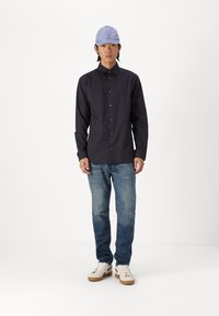 Camisa de rayas oscura de manga larga, con cierre de botones y gorra azul. Lleva puesta unos jeans de mezclilla azul claro y zapatillas blancas con suelas beige.