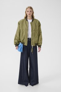 Πράσινο-ελιάς bomber μπουφάν με ραμμένες λεπτομέρειες, συνδυασμένο με φαρδιά σκούρα τζιν παντελόνια και μικρή μπλε τσάντα χειρός.