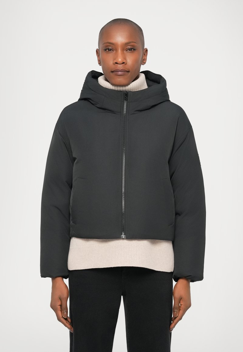 Chaqueta puffa corta negra con capucha, que presenta un cierre frontal y bolsillos laterales. Lleva puesta sobre un suéter de punto crema, con pantalones negros de corte holgado.