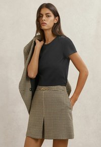 Juoda trumpomis rankovėmis marškinėliai derinami su rudai juodu žirnio danties rašto sijonu, kuriame yra klostė ir šoniniai kišenės.