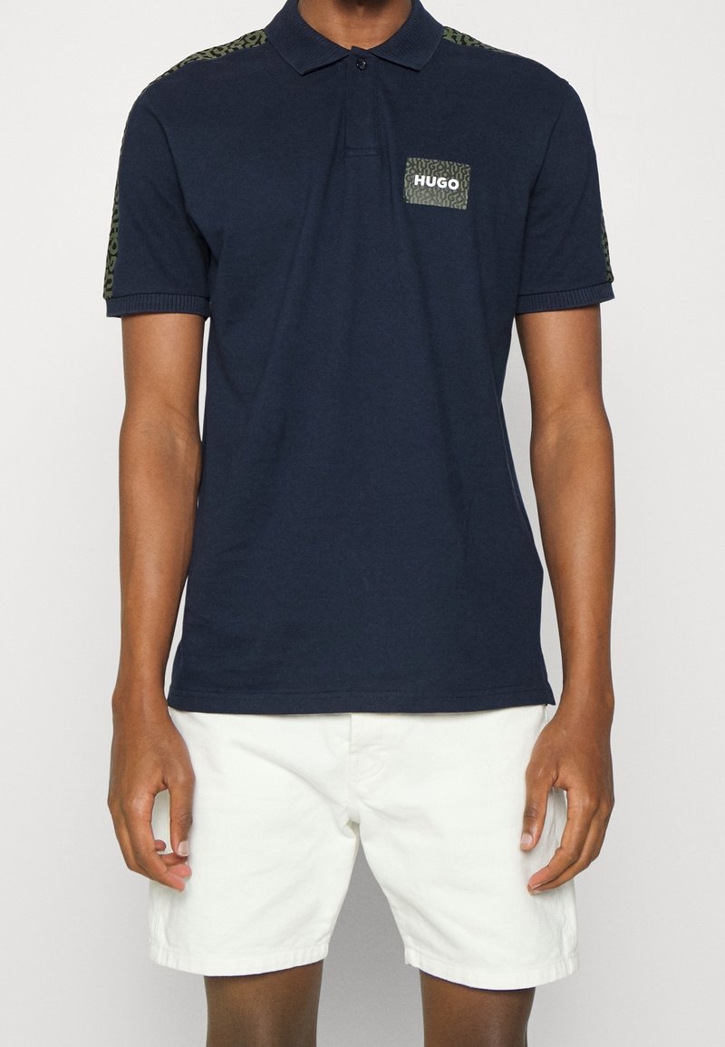 Polo bleu marine avec un accent texturé à motif léopard vert sur l'épaule et un patch de marque sur la poitrine. Manches courtes, col classique.