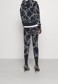 Marinträningsdräkt i marinblått med ett vitt kedjemönster, inklusive en huvad sweatshirt och tapered byxor. Kombinerad med svarta högklackade skor.