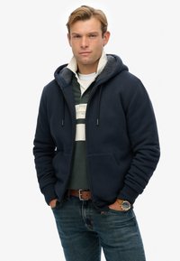 Marineblå zip-hoodie med en blød indvendig foring, designet med to forlommer, båret over en grøn og hvidstribet skjorte. Slidte denim jeans.