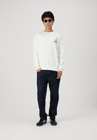 Balta sporta krekla ar trīsstūra grafiku uz krūšu daļas, apvienota ar tumši ziliem džinsiem un melniem sporta apaviem. Modelis valkā tumšas saulesbrilles.