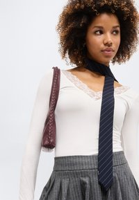 Junge Frau mit lockigem Haar trägt ein weißes, spitzenbesetztes Oberteil, einen grauen Nadelstreifenrock, eine marineblaue gestreifte Krawatte und eine kastanienbraune Leder-Umhängetasche.