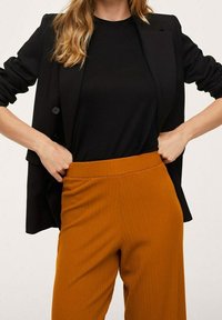 Blazer noir sur un haut noir, associé à un pantalon orange côtelé taille haute. Tissu lisse, col rond, coupe décontractée.