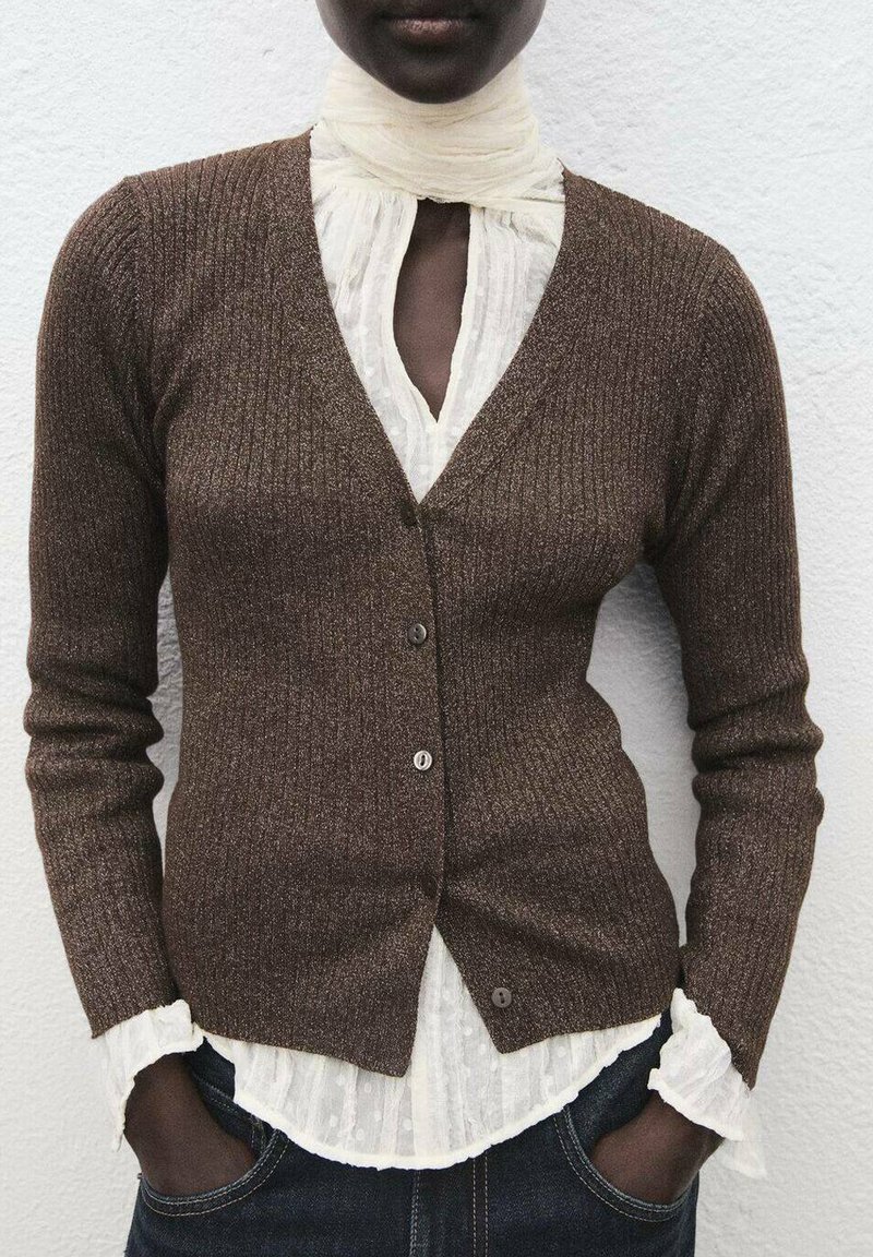 Hnědý žebrovaný cardigan s V-šichem, těsným střihem a pěti knoflíky. Nositý přes bílou texturovanou blůzu s volánovými manžetami a límečkem.