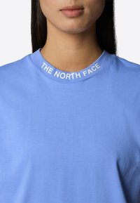 Světle modré tričko s kulatým výstřihem, na límci je bílé text "THE NORTH FACE". Měkká, hladká textura látky.