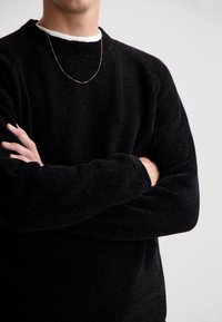 Schwarzer Strickpullover mit strukturiertem Muster und geripptem Kragen, über einem weißen Hemd getragen. Eine dünne goldene Kette ist um den Hals sichtbar.