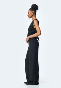 Svart jumpsuit med en åtsittande bodice, breda ben och v-ringning. Har knäppning och ett knyt i midjan. Slät, strukturerad tyg.