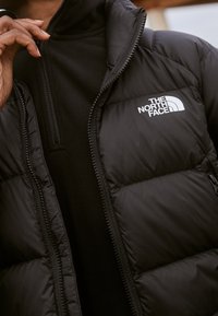 Veste noire rembourrée avec fermeture éclair, dotée d'une doublure en polaire et d'un logo blanc brodé sur la poitrine gauche. Design texturé et matelassé.