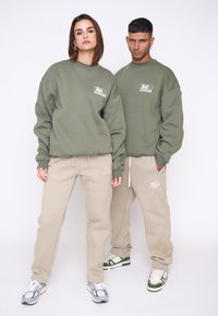 Unisex oversized sweatshirt in olijfgroen met logo; beige joggingsbroek met borduursel. Beide outfits hebben een ontspannen pasvorm en zachte stof.