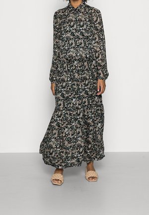 Kobieta ubrana w długą, czarną sukienkę w kwiaty z długimi rękawami i zapinanym na guziki kołnierzykiem, w połączeniu z beżowymi plecionymi sandałami wsuwanymi.