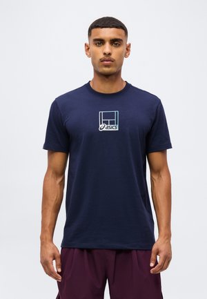 Homme portant un t-shirt bleu marine Asics avec un petit logo sur la poitrine et un short marron, debout devant un fond clair uni.