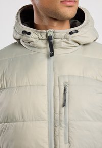 Chaqueta aislante gris claro con capucha, que cuenta con un cierre frontal de cremallera, tejido texturizado y un bolsillo en el pecho con cremallera.