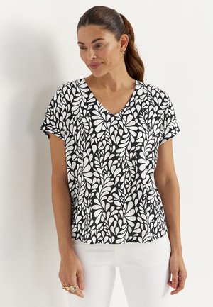 Kobieta o ciemnych włosach związanych w kucyk, ubrana w czarno-biały wzorzysty top z krótkim rękawem i dekoltem w serek oraz białe spodnie, stojąca na białym tle.