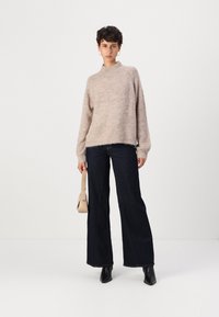 Light brown knitted sweater with a high neck, paired with dark wide-leg jeans. Model holds a small beige bag and wears black ankle boots.