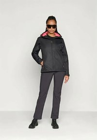 Femme portant une veste noire avec une doublure rose, un pantalon foncé, des bottes noires et de grandes lunettes de soleil noires, se tenant devant un fond blanc uni.