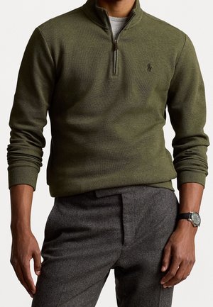 Mann trägt olivgrünen Pullover mit Reißverschluss bis zur Brust über grauer Hose, mit schwarzer Armbanduhr am linken Handgelenk, steht vor einfachem Hintergrund.