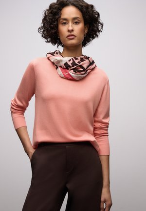 Nainen, jolla on kiharat lyhyet hiukset, pukeutunut vaaleanpunaiseen pitkähihainen paita, mustat housut ja kaulassaan kuvioitu huivi, poseeraa yhdellä kädellä taskussa.