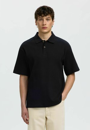 Jeune homme aux cheveux courts et bouclés portant un polo noir côtelé à manches courtes et un pantalon beige, debout devant un fond blanc uni.