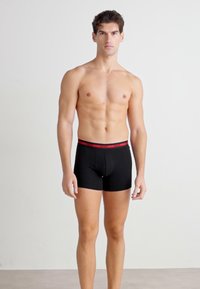 Boxers noirs avec une ceinture rouge, fabriqués à partir d'un tissu lisse et extensible, avec un design ajusté et une poche contour.
