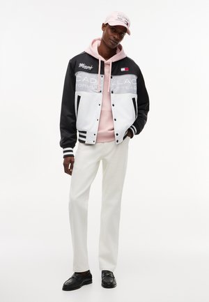Ψηλός άνδρας που φοράει μαύρο και λευκό μπουφάν αγώνων, ροζ φούτερ με κουκούλα και καπέλο, λευκό παντελόνι και μαύρα μοκασίνια, στέκεται μπροστά σε απλό λευκό φόντο.