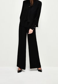 Femme portant un chemisier noir à manches longues avec des boutons dorés et un pantalon noir à jambes larges, debout sur un fond blanc uni.