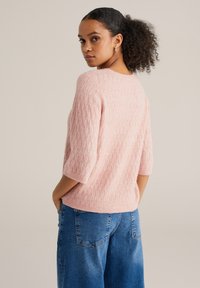 Pulover roz tricotat cu model texturat în formă de diamant, mâneci scurte și croială lejeră, asortat cu blugi albaștri cu picioare largi.