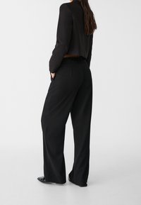 Vrouw in zwarte wijde broek en cropped zwarte blazer, staand met handen in de zakken, met de rug naar de camera op een plain witte achtergrond.