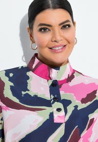 Blouse colorée avec un col haut, présentant un motif abstrait en rose, vert, bleu marine et bordeaux. Trois boutons à l'avant.