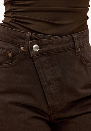 Pantalon en velours côtelé marron à coupe ajustée, avec un bouton métallique, des passants pour ceinture et une texture subtile. Poches visibles de chaque côté.