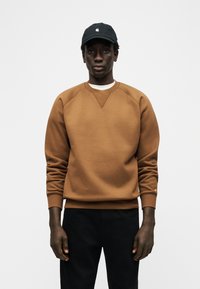 Άνδρας που φοράει καφέ φούτερ, μαύρο καπέλο και μαύρο παντελόνι, στέκεται μπροστά σε λευκό φόντο, κοιτάζοντας μπροστά.