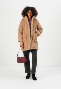 Beige Fleece-Mantel mit Kapuze, großen Taschen und Knopfverschluss. Kombiniert mit dunkelgrauen Hosen und schwarzen Stiefeletten, hält sie eine burgunderfarbene Handtasche.